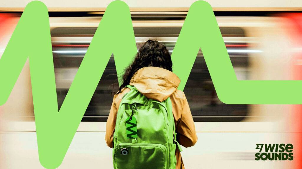Una persona con una mochila verde espera en una estación de metro mientras se desplaza un tren en el fondo.