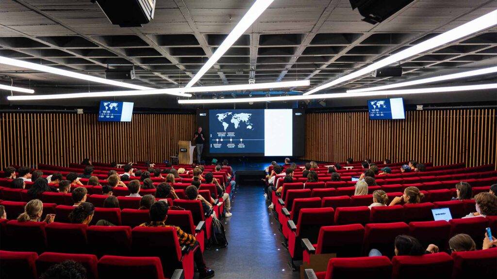 Una conferencia en un auditorio con numerosas personas sentadas en filas rojas.