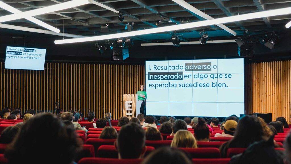 Una conferencia con un ponente frente a una pantalla que muestra texto en una sala llena de público.