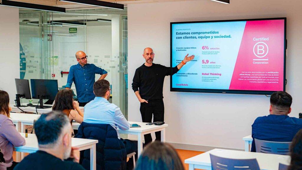 Un grupo de personas asiste a una presentación en un aula moderna con pantallas y mesas de trabajo.