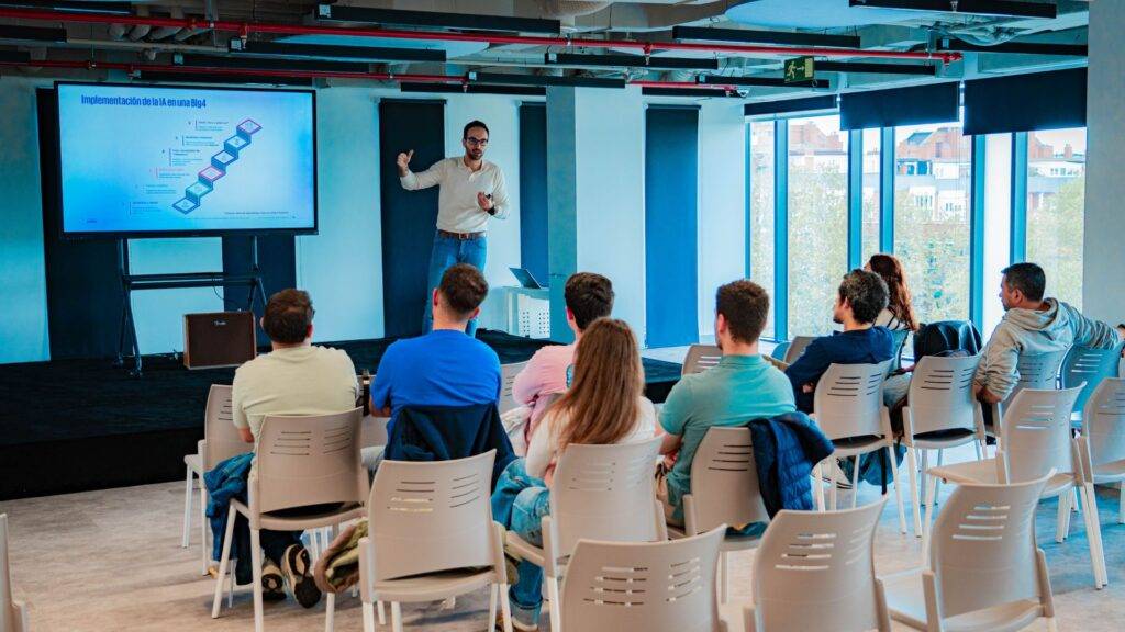 Un presentador expone ante un grupo de personas en una sala moderna con pantallas y ventanas grandes.