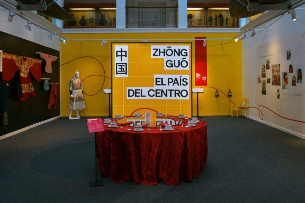 La imagen muestra una exposición cultural con un fondo amarillo y objetos de arte en un ambiente de museo.