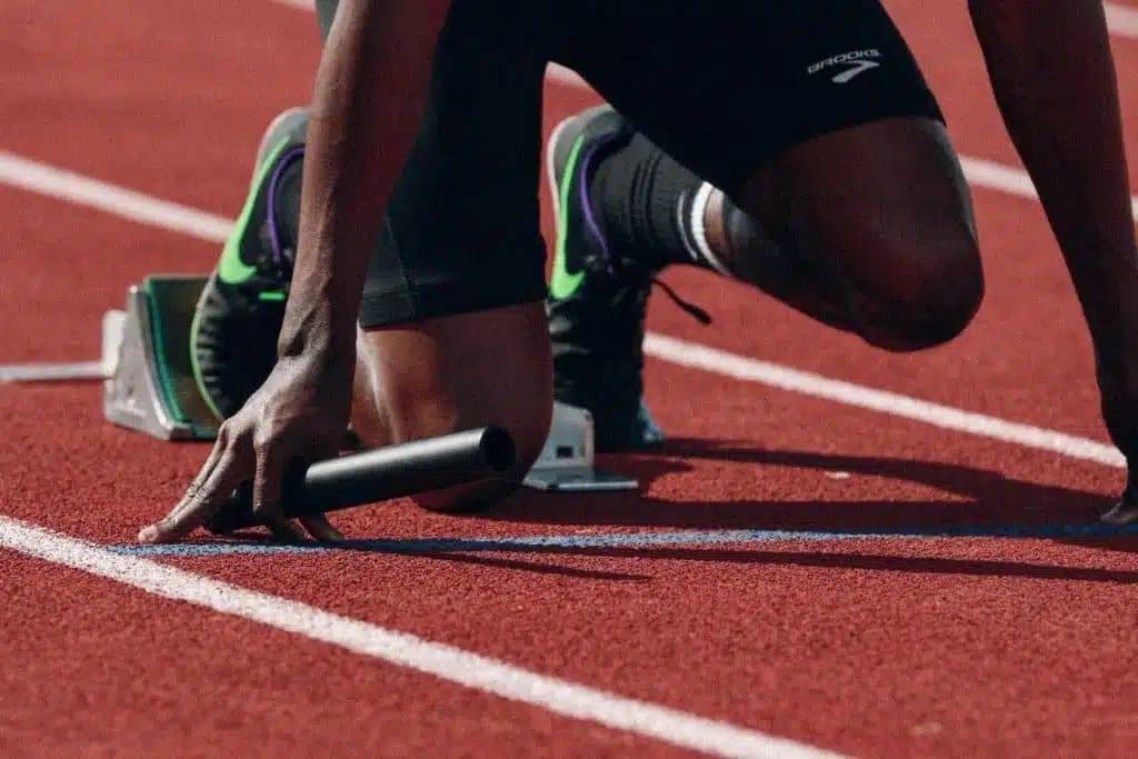 Un atleta está preparado en la pista de atletismo, con una mano en el suelo y sosteniendo un testigo de relevo.