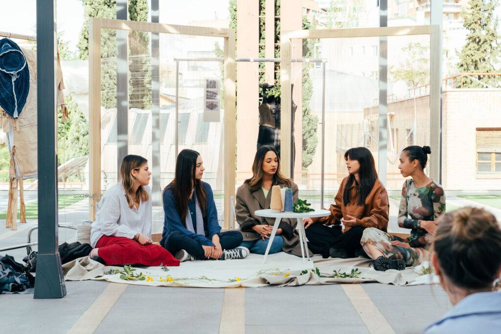 Un grupo de mujeres se sienta en círculo en un ambiente al aire libre, conversando alrededor de una mesa pequeña.