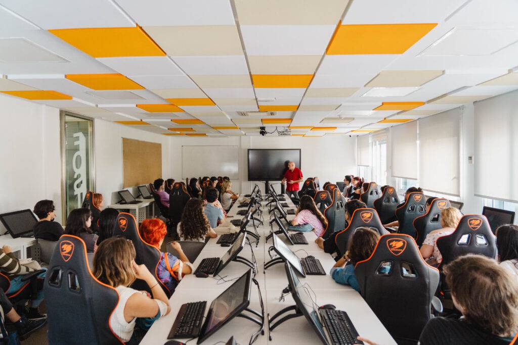 Una sala de aula con muchos estudiantes sentados frente a computadoras mientras un profesor da una charla.