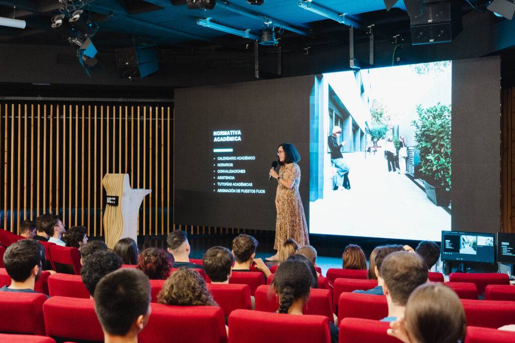 Una presentadora habla frente a una audiencia en un ambiente académico mientras se proyecta una imagen en la pantalla.