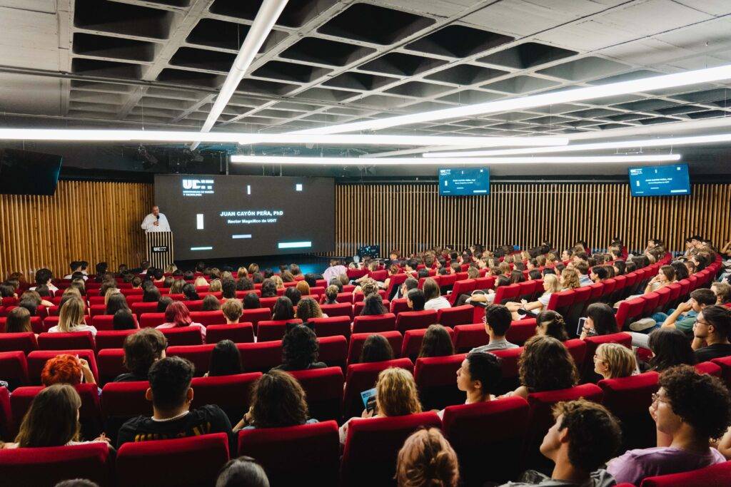 Auditorio con un gran número de personas sentadas en filas rojas, escuchando a un orador en el escenario.