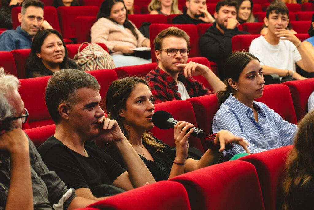 Un grupo de personas en un auditorio, escuchando a una mujer que está haciendo una pregunta con un micrófono en mano.