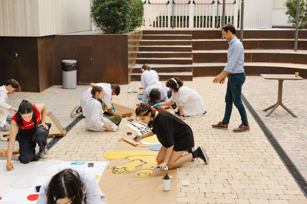 Un grupo de personas trabaja en una actividad artística al aire libre, pintando sobre cartones en el suelo.