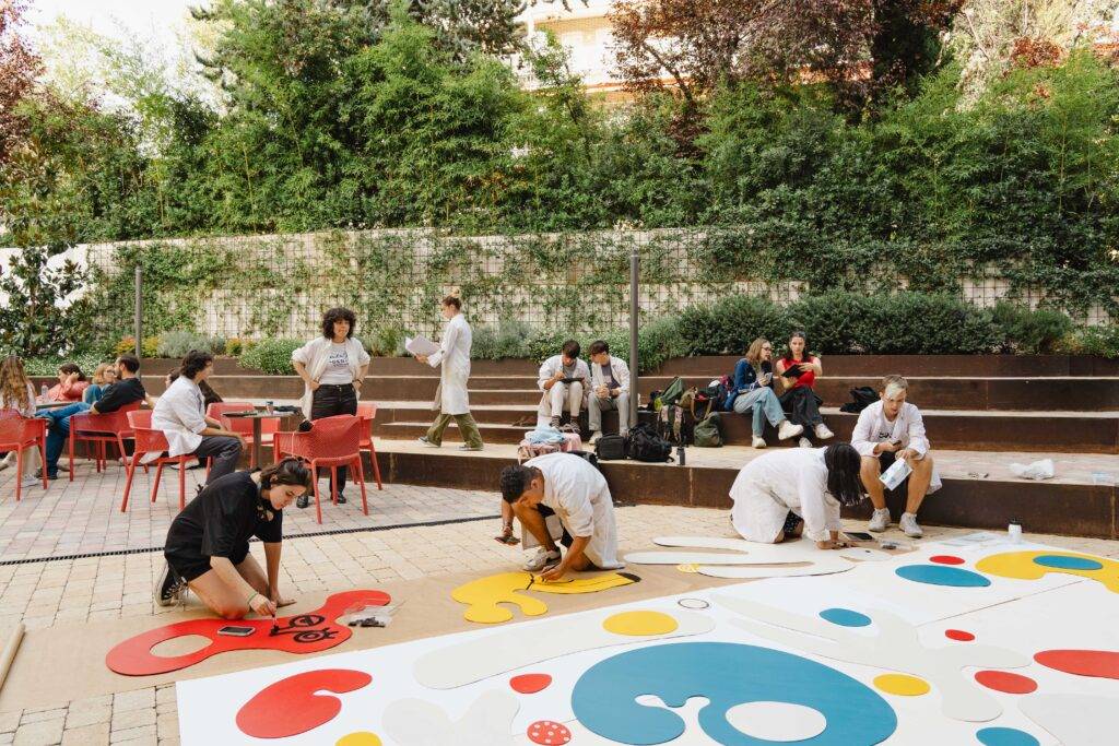 Un grupo de personas trabaja en un mural colorido en un espacio al aire libre mientras otros observan y disfrutan del ambiente.