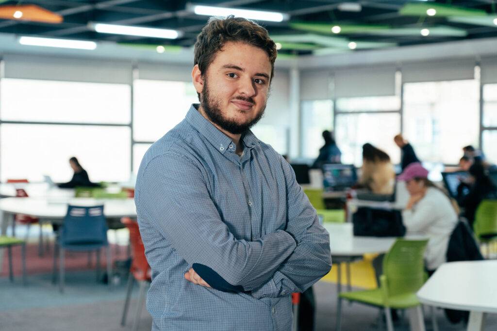 Un joven sonriente se presenta en un ambiente de estudio moderno con varias personas al fondo.