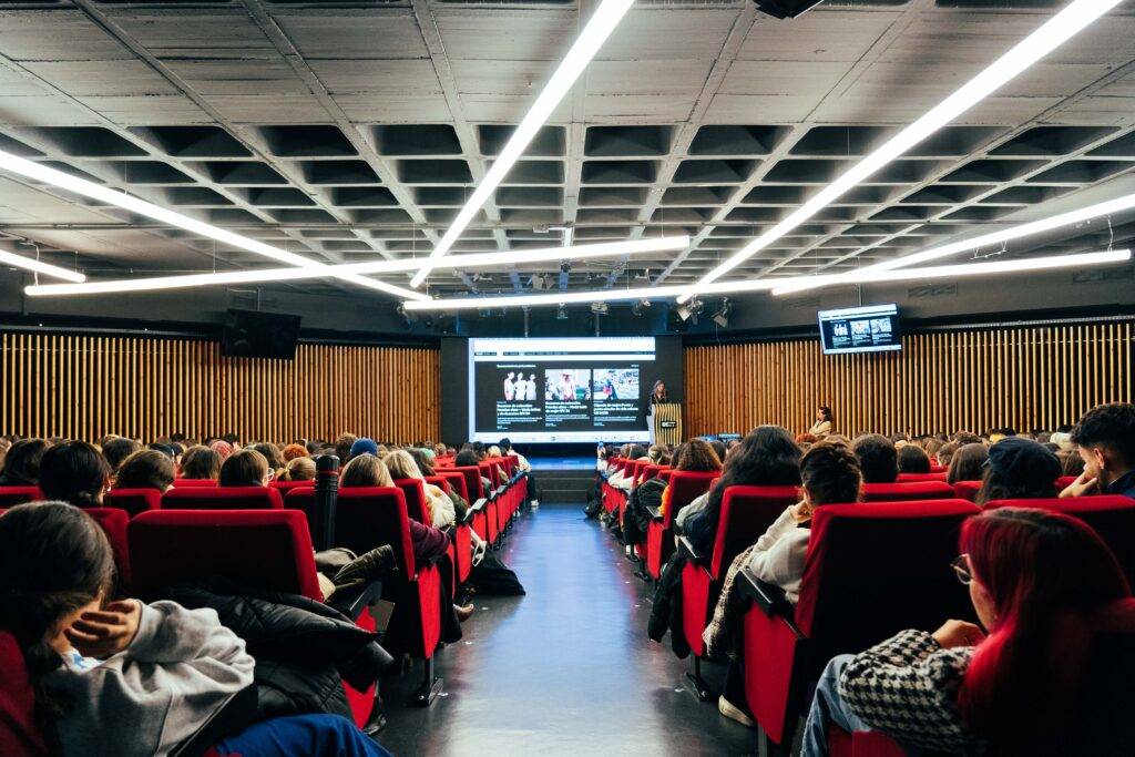 Una sala con muchas personas sentadas en sillones rojos, mirando una presentación en una pantalla.