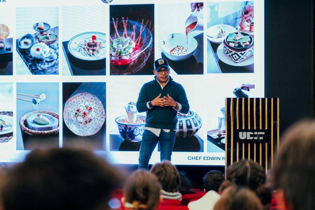 Un chef presenta su trabajo culinario mientras muestra una serie de platos en una pantalla detrás de él.