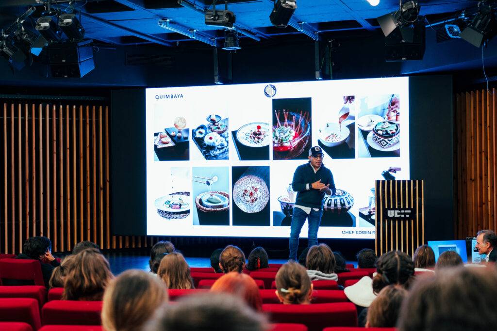 Un chef presenta platos en una conferencia con imágenes en una pantalla detrás de él.