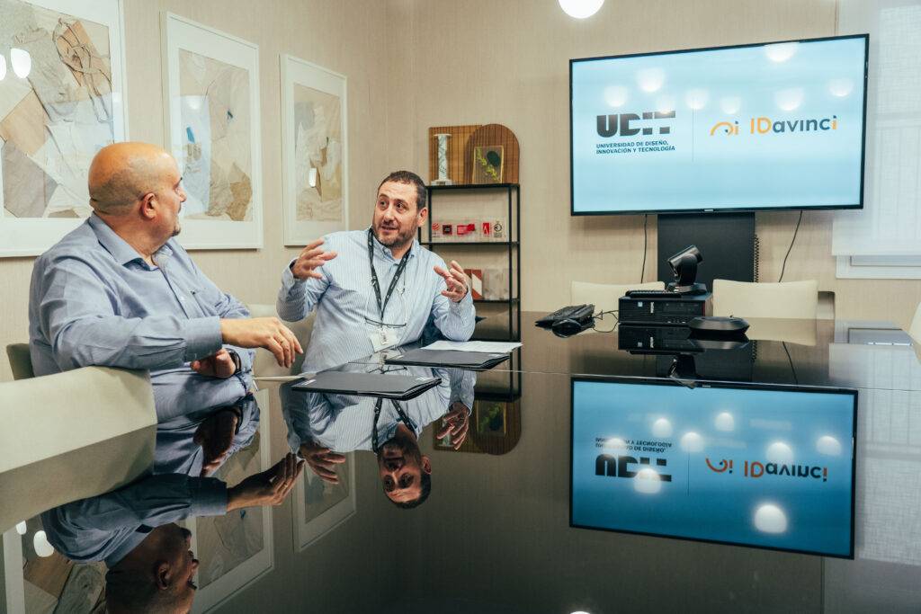 Dos hombres conversan animadamente en una sala de juntas con una pantalla al fondo.