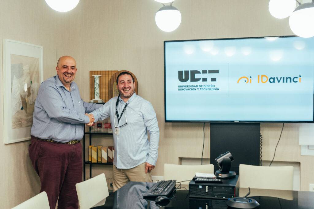 Dos hombres se dan la mano en una reunión junto a una pantalla que muestra los logotipos de la universidad y una empresa.