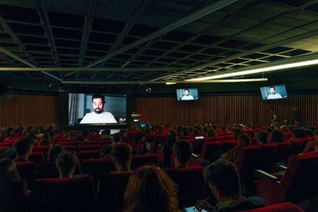 Una sala de conferencias con un presentador en una pantalla y un público sentado en asientos rojos.