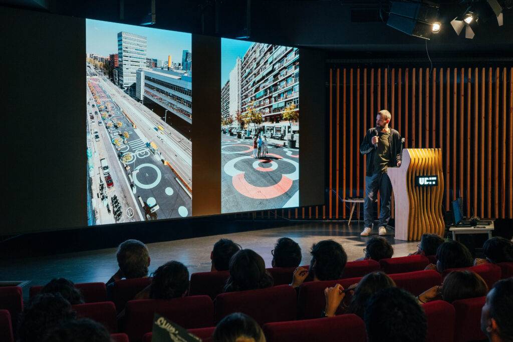 Un orador presenta imágenes de un diseño urbano en un auditorio lleno de gente.