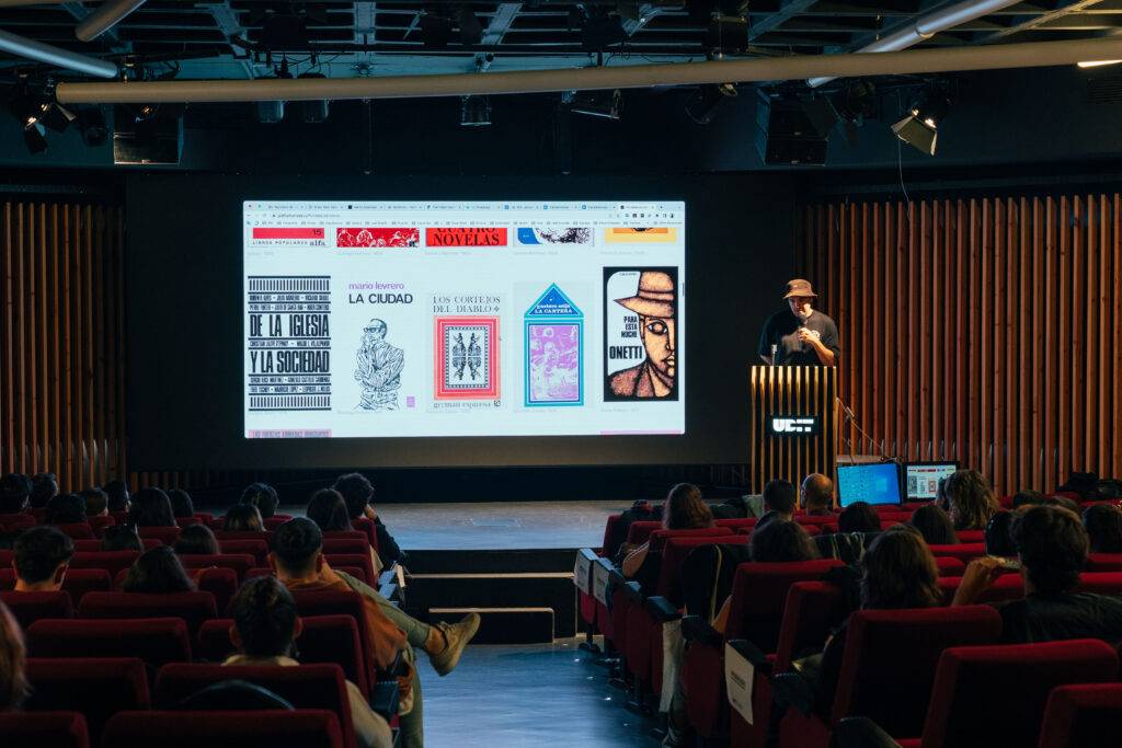 Un presentador en un escenario habla ante una audiencia mientras se proyectan imágenes de libros en una pantalla.