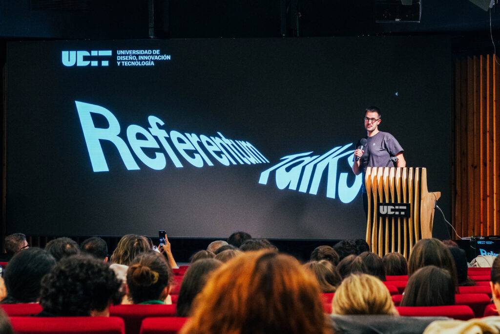 Un ponente presenta en una conferencia en una sala llena de gente con un fondo de proyección donde se lee 'Referentium'.