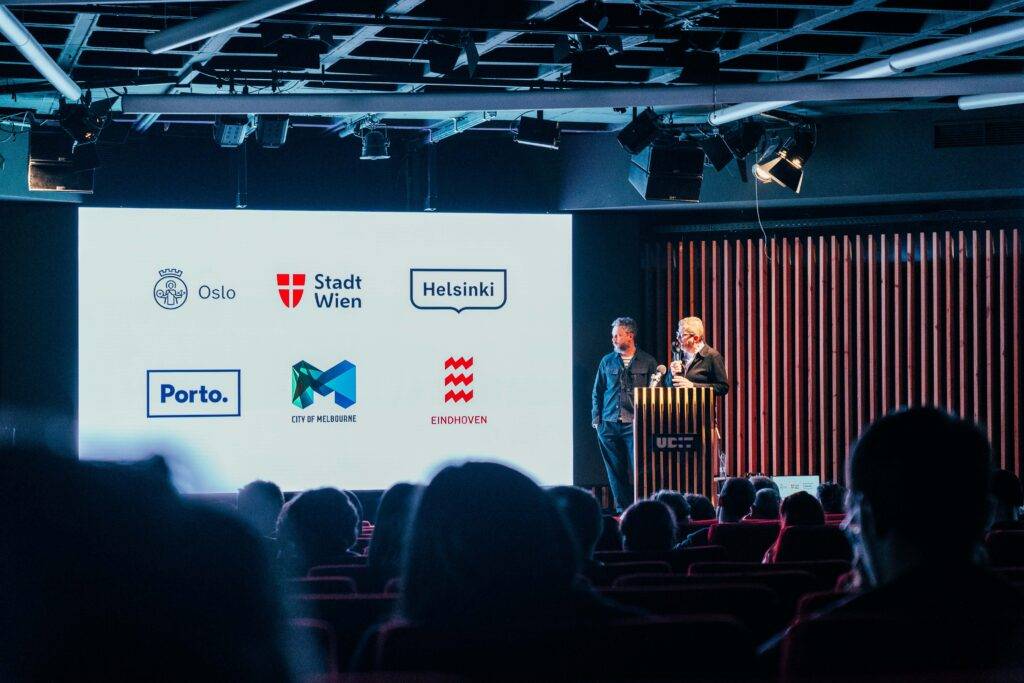 Un evento donde dos presentadores están hablando frente a una audiencia, con un proyector que muestra varios logotipos de ciudades.