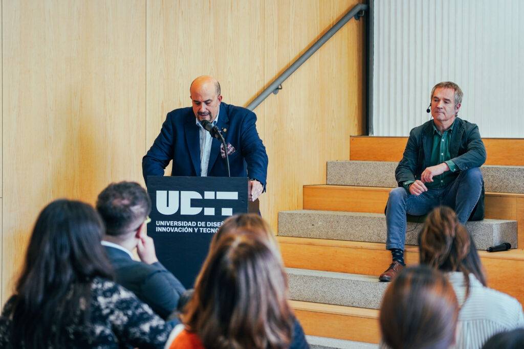 Un orador presenta en un evento en la Universidad de Diseño, mientras un panelista escucha desde el escenario.