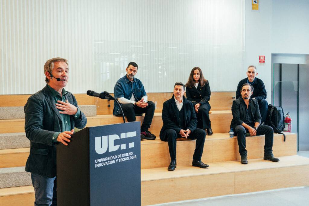 Un grupo de personas sentadas en un escenario mientras un hombre habla desde un micrófono.
