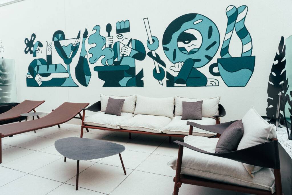 La imagen muestra un mural colorido en un espacio al aire libre con sillones y mesas modernas.