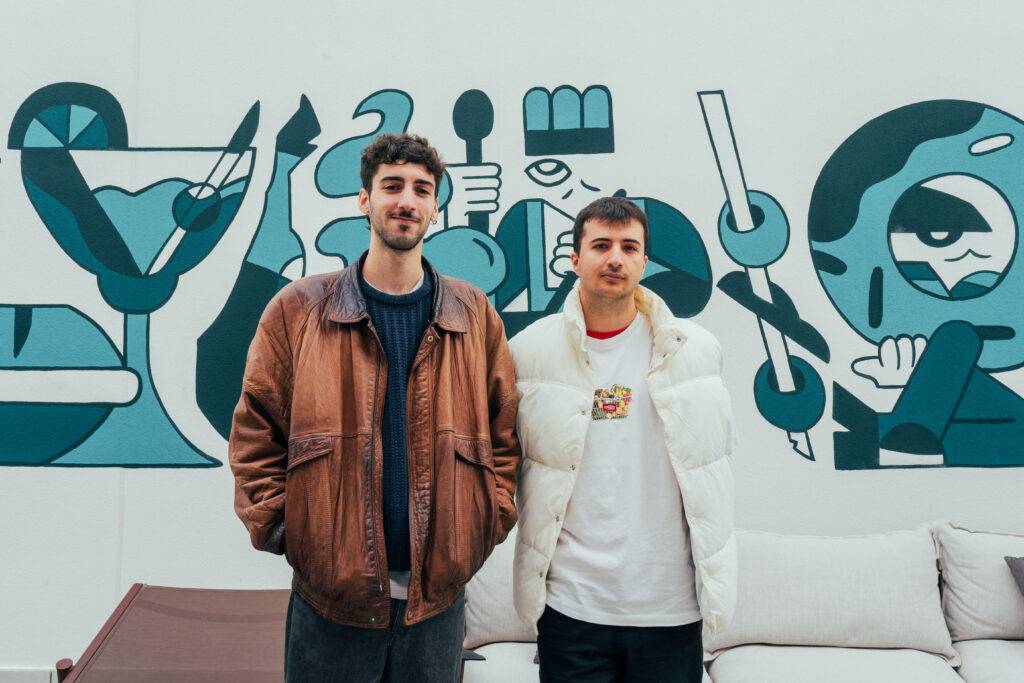 Dos jóvenes posan juntos frente a un mural colorido en un ambiente moderno.