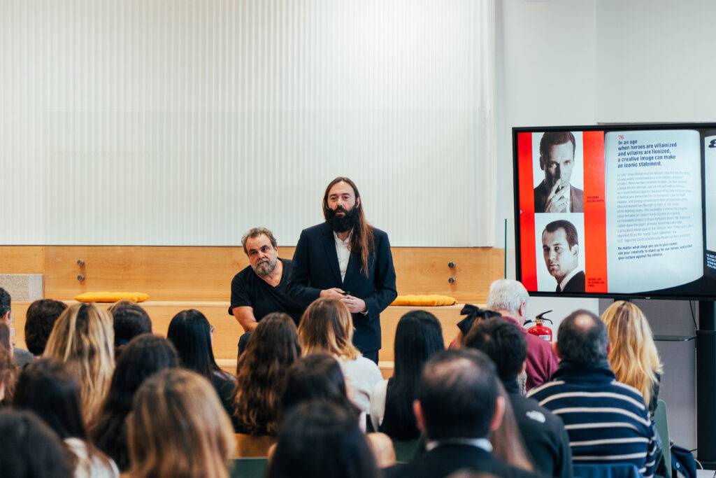 Una presentación en un auditorio donde un hombre con barba y otro con cabello corto están hablando frente a una audiencia.