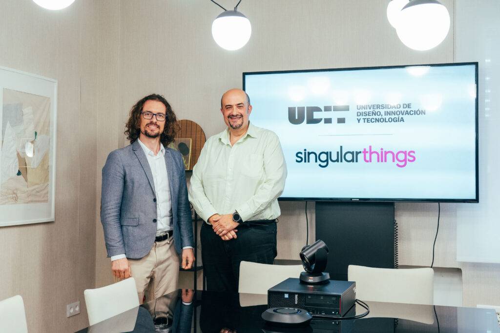 Dos hombres sonrientes posan en una sala de reuniones con un logo visible de una universidad y una empresa.