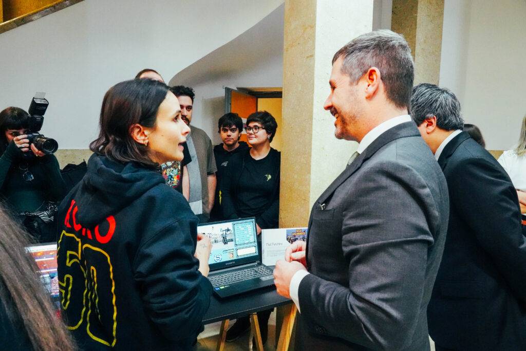 Una joven con sudadera negra conversa amigablemente con un hombre trajeado en un evento, mientras un grupo de personas observa al fondo.
