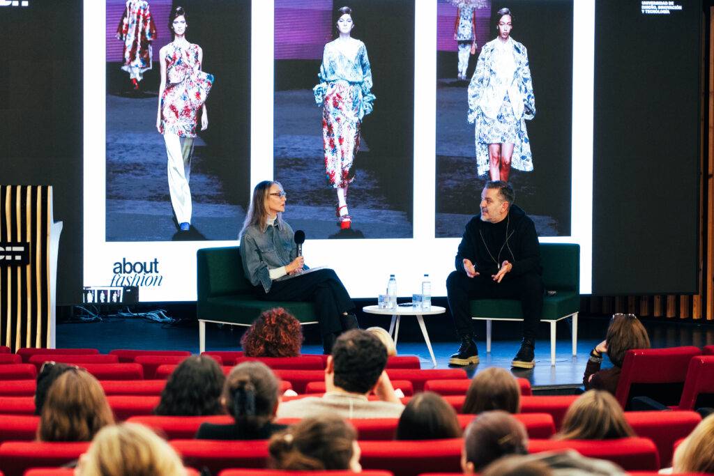 Un evento de moda con dos oradores en un panel, y una pantalla detrás mostrando tendencias de vestimenta.