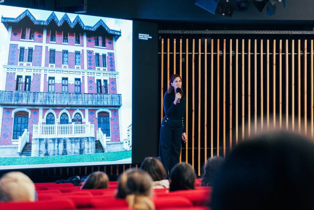 Una mujer está hablando frente a una audiencia, con una imagen de un edificio colorido proyectada detrás.