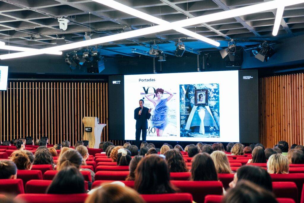 Una conferencia donde un ponente presenta revistas en una pantalla frente a un auditorio lleno de personas sentadas.