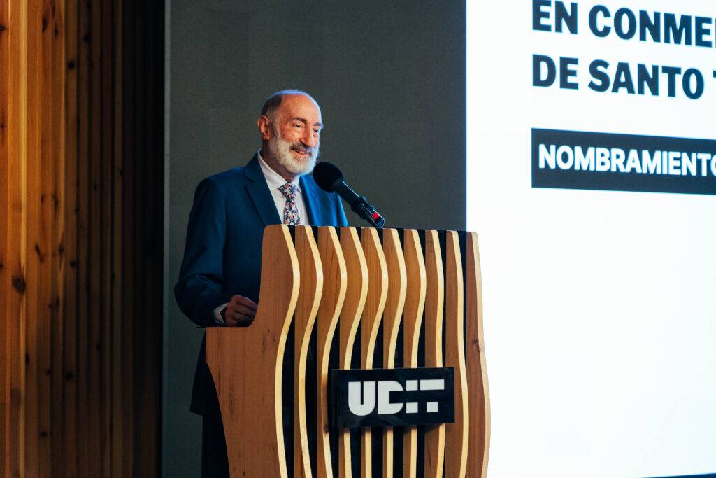 Un hombre de pie detrás de un podio presenta un evento con un cartel detrás que menciona un nombramiento.