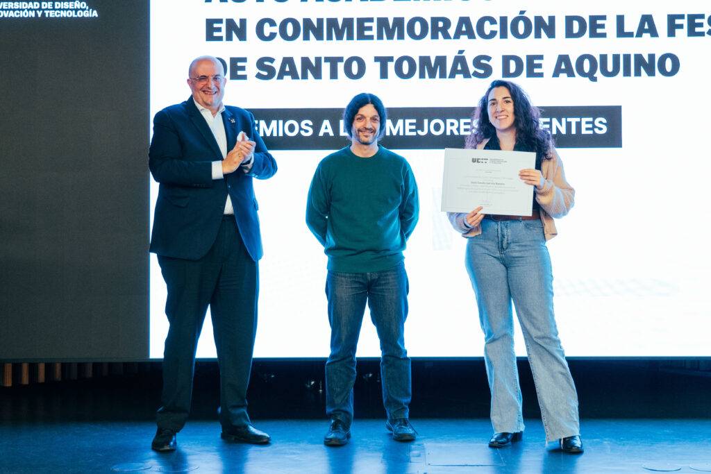 Una ceremonia de premiación donde tres personas están de pie frente a un fondo que incluye un texto sobre la festividad de Santo Tomás de Aquino.