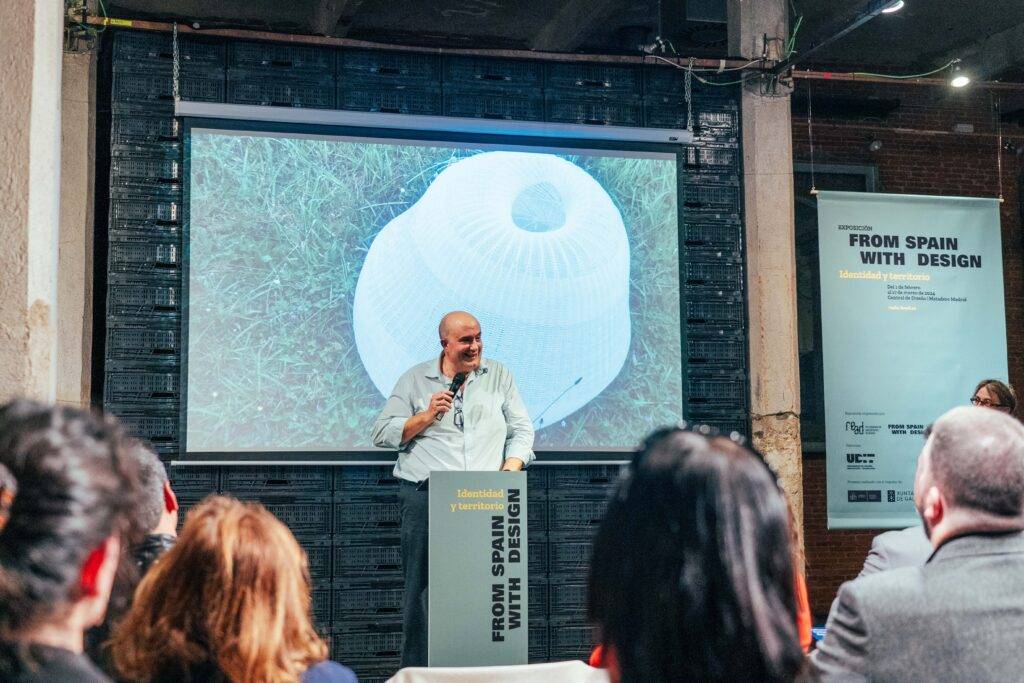 Un hombre habla en un evento sobre diseño, mientras una proyección de un objeto moderno se muestra en la pantalla detrás de él.