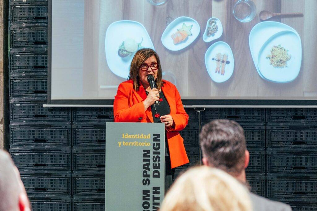 Una mujer con un abrigo naranja presenta frente a una audiencia mientras muestra un diseño gastronómico en una pantalla.