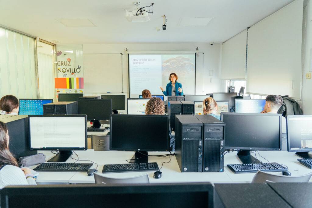 Una profesora da una clase en un aula con varios estudiantes sentados frente a computadoras.