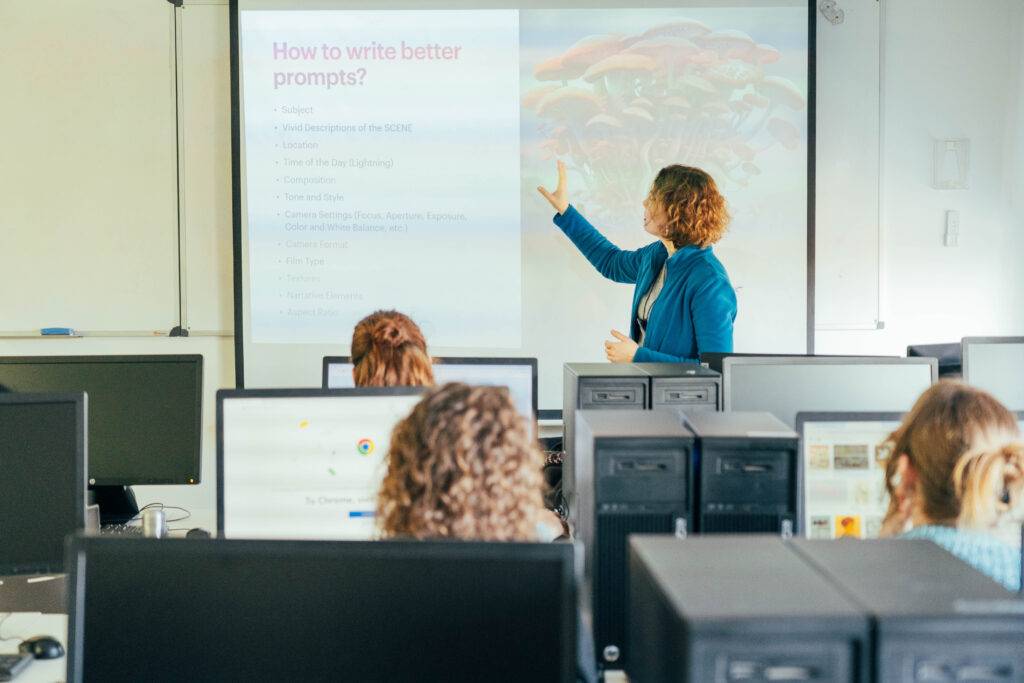 Una profesora está enseñando sobre cómo redactar mejores indicaciones en una clase llena de estudiantes frente a computadoras.