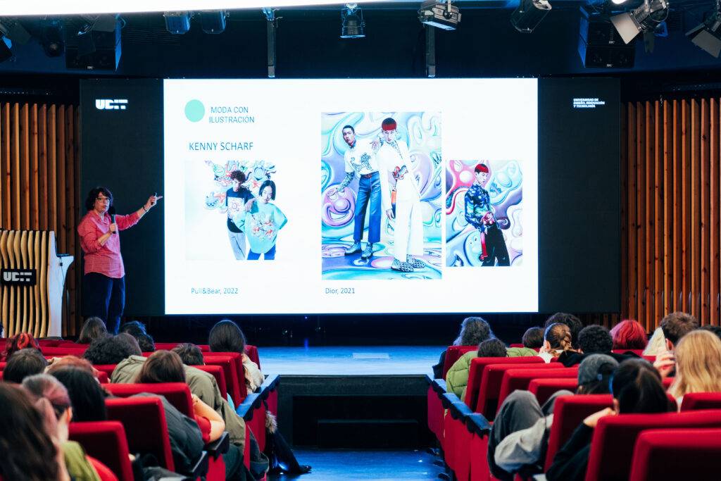 Una presentadora muestra una diapositiva sobre el trabajo de Kenny Scharf en una conferencia llena de espectadores.