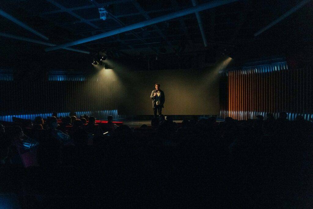 Un orador se presenta en el escenario frente a una audiencia en un ambiente iluminado débilmente.
