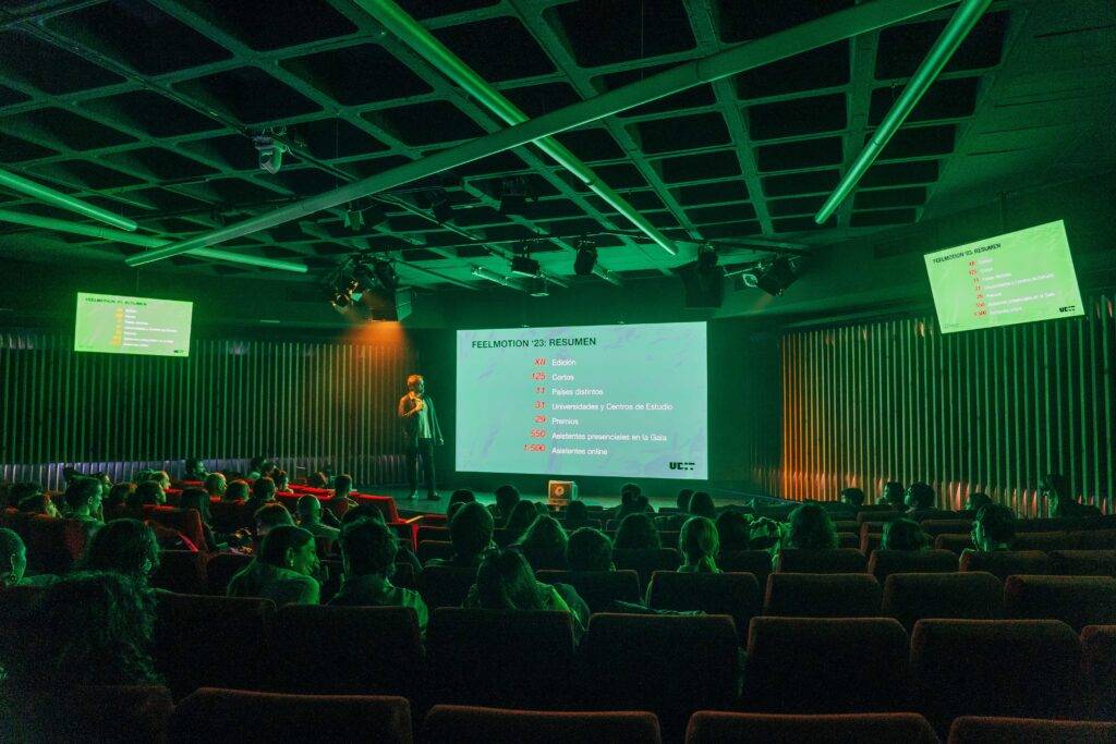 Una presentación en un auditorio iluminado de verde, donde un ponente está hablando frente a una audiencia sentada.