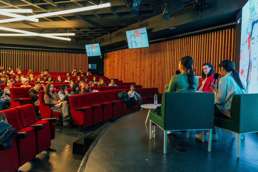 Una charla o evento en un auditorio moderno con público atento y panelistas en el escenario.