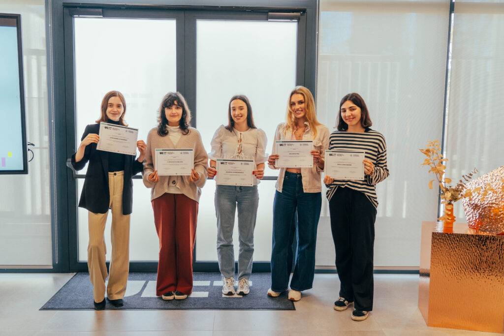 Un grupo de cinco mujeres sonrientes sostiene certificados frente a una pared de vidrio.