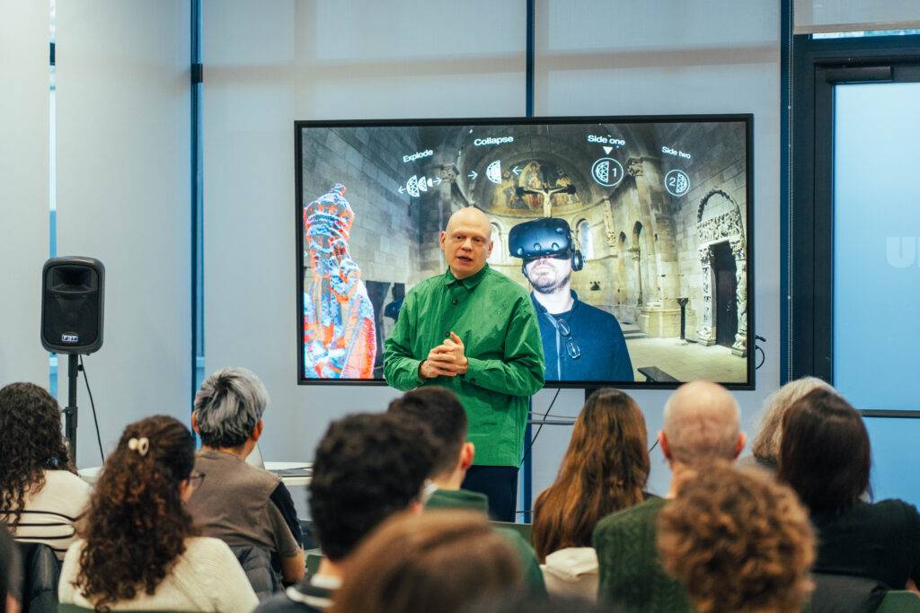 Un ponente presenta una charla sobre tecnología en una sala con un proyector mostrando imágenes detrás de él.