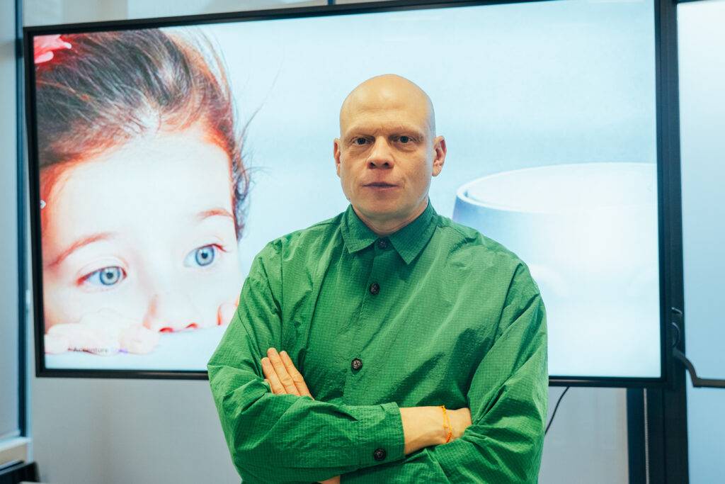 Un hombre calvo vestido con una camisa verde se encuentra frente a una pantalla que muestra la imagen de una niña con mirada curiosa.
