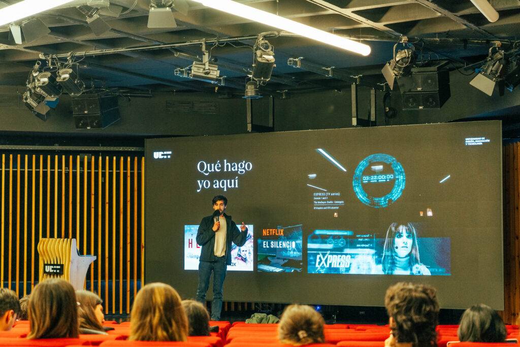 Un presentador está hablando frente a una pantalla grande en un auditorio con el título 'Qué hago yo aquí'.
