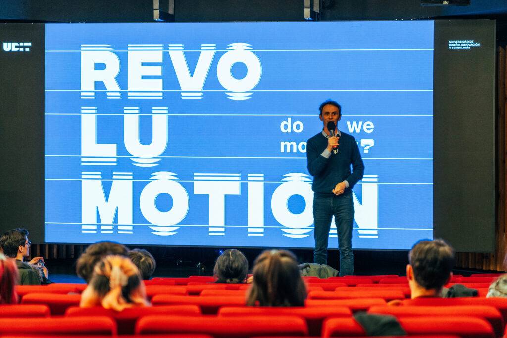 Un presentador está hablando frente a una gran pantalla con el texto 'REVOLU MOTION'.